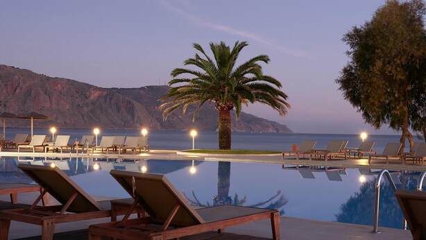 Zona junto a la piscina con sillones de descanso y una palmera, con vistas a un cuerpo de agua y montañas.