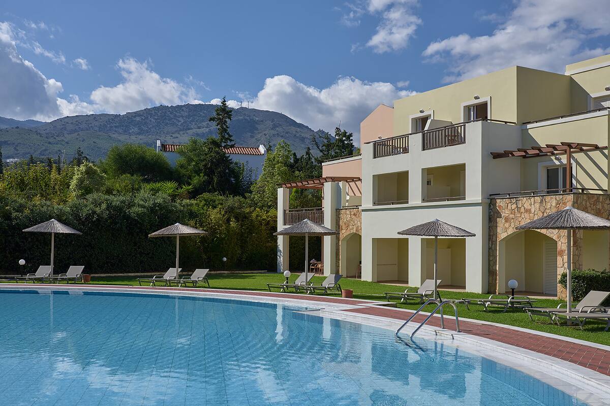 Piscina con sillas de descanso y sombrillas, rodeada por un edificio de fachada de piedra y una montaña al fondo.