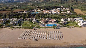 Vista aérea de un complejo turístico costero con varios edificios, una piscina grande y una playa con numerosas tumbonas.