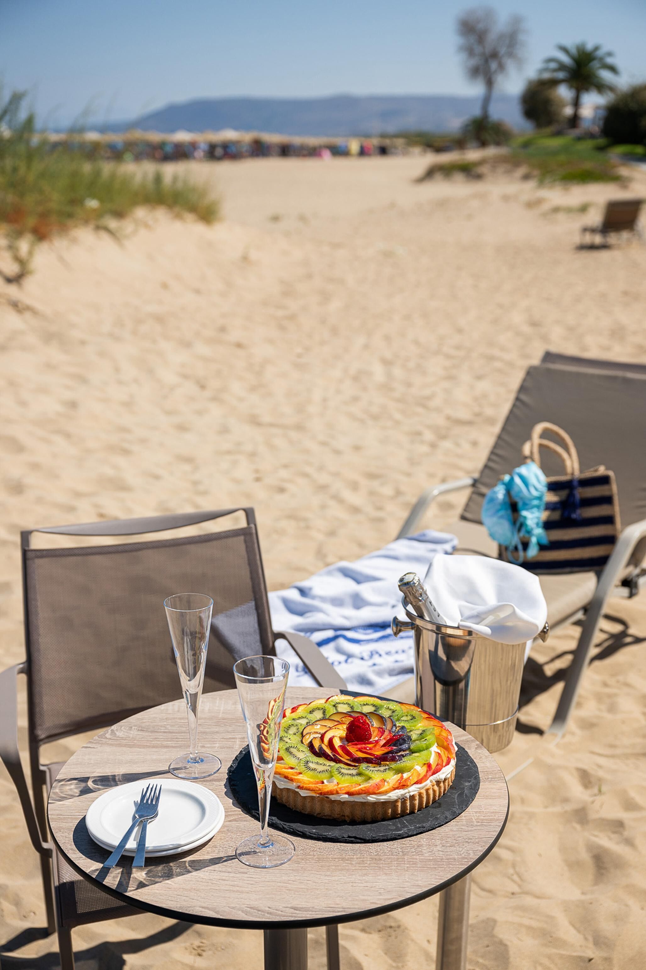 A beachside table with a dessert, a glass of champagne, and a bottle of water, set against a backdrop of a sandy beach and lounge chairs.
