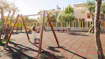 A playground with swings and a slide, surrounded by a stone wall and trees.