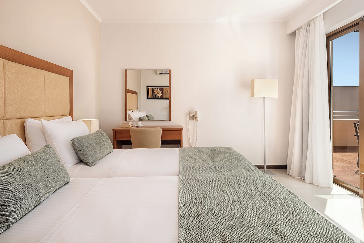 A hotel room with a bed, a desk, a chair, a lamp, a mirror, and a view of the outside.