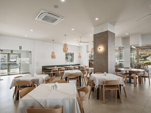 A spacious dining area with multiple tables and chairs, a large pillar, and a view of the outdoors. - Sirios Village Hotel & Bungalows - All Inclusive (Chania)