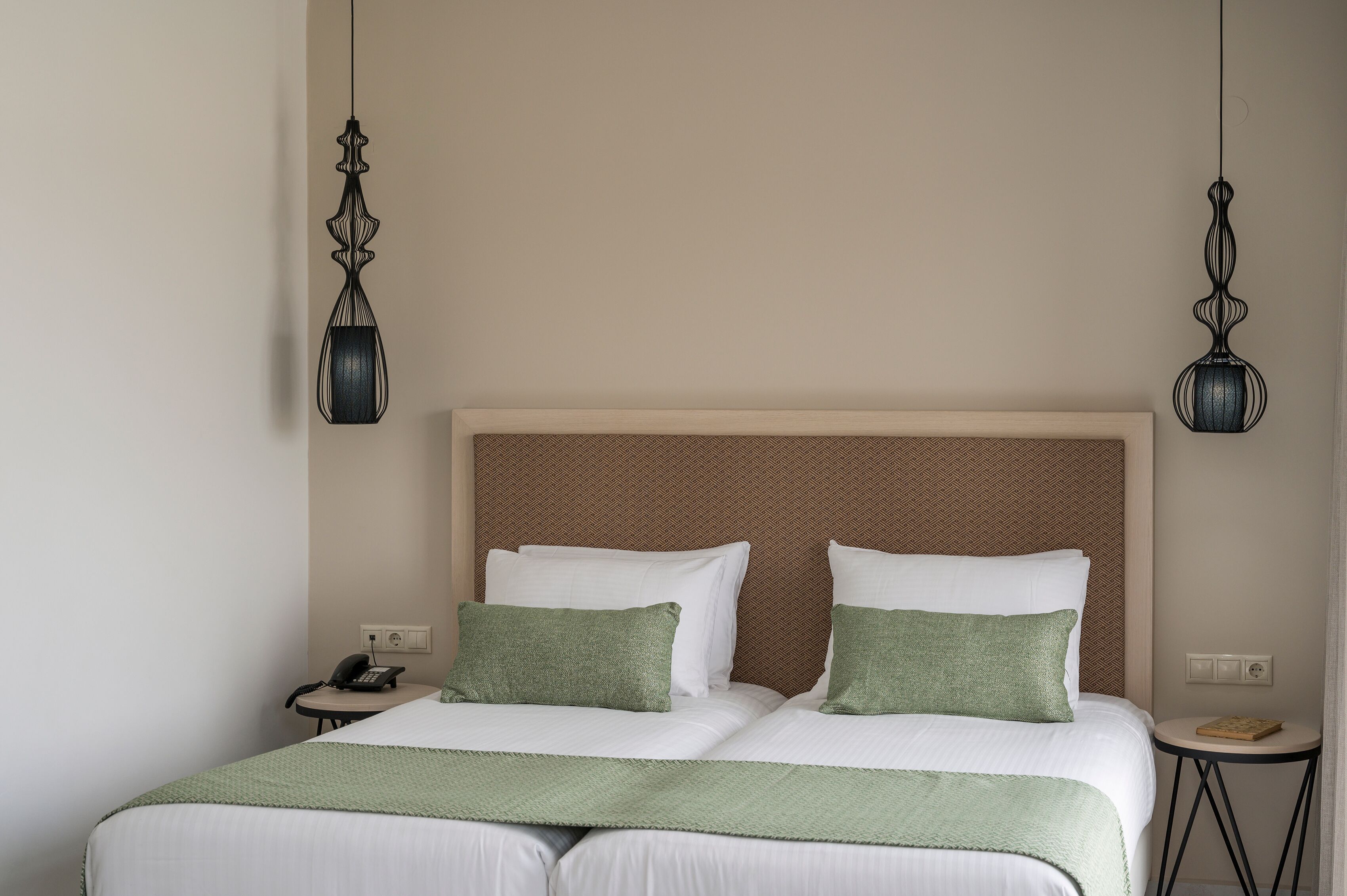 A double bed with green pillows, a wooden headboard, a bedside table with a phone, and two hanging lamps.