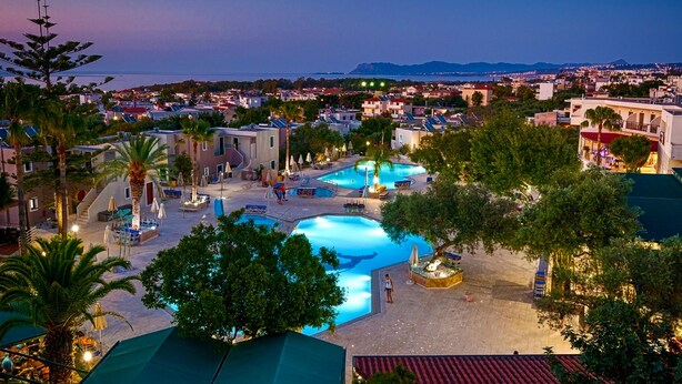 Un resort con varias piscinas, palmeras y vistas al mar y montañas distantes.