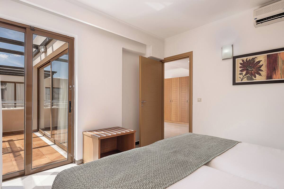A modern bedroom with a large bed, a small wooden bench, a sliding glass door leading to a balcony, and a wooden wardrobe.