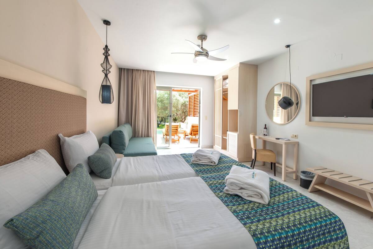 A hotel room with two beds, a desk, a chair, a ceiling fan, and a balcony with outdoor seating.