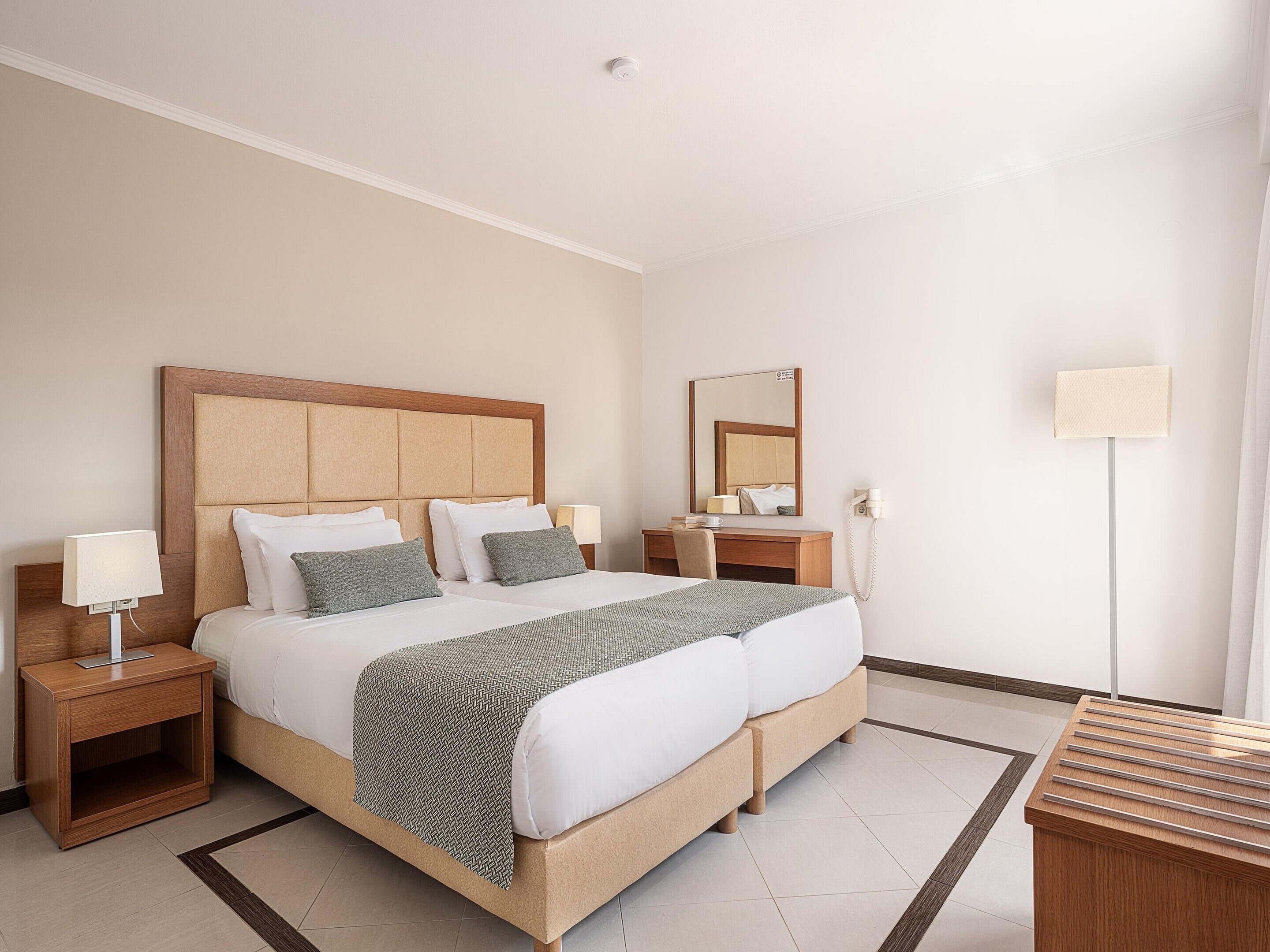 A hotel room with a bed, bedside tables, a desk, a chair, a mirror, and a lamp.
