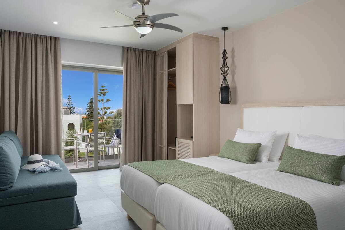 A hotel room with a large bed, a ceiling fan, a balcony with a table and chairs, and a view of trees and buildings.