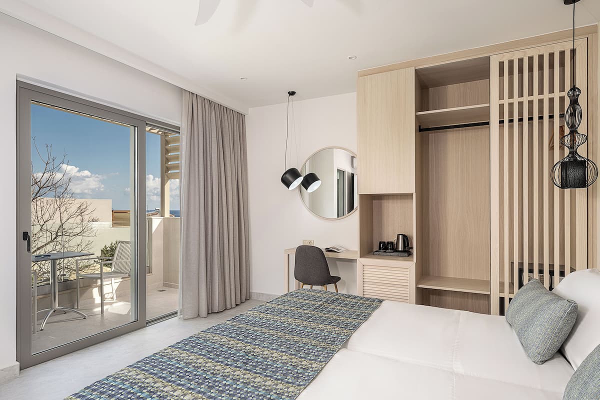 A modern hotel room with a large bed, a desk, a chair, a wardrobe, and a balcony with a view.