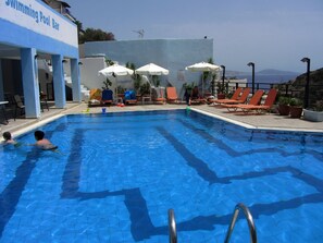 A swimming pool with lounge chairs and umbrellas, a bar, and a view of the sea. - Petra Hotel (Agios Vasileios)