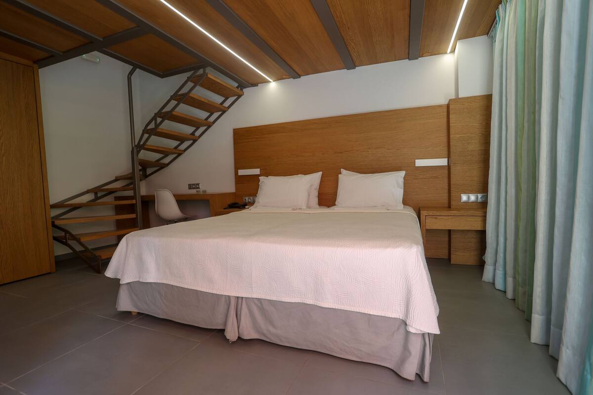 A modern bedroom with a large bed, wooden headboard, and a built-in desk. A staircase is visible to the left.