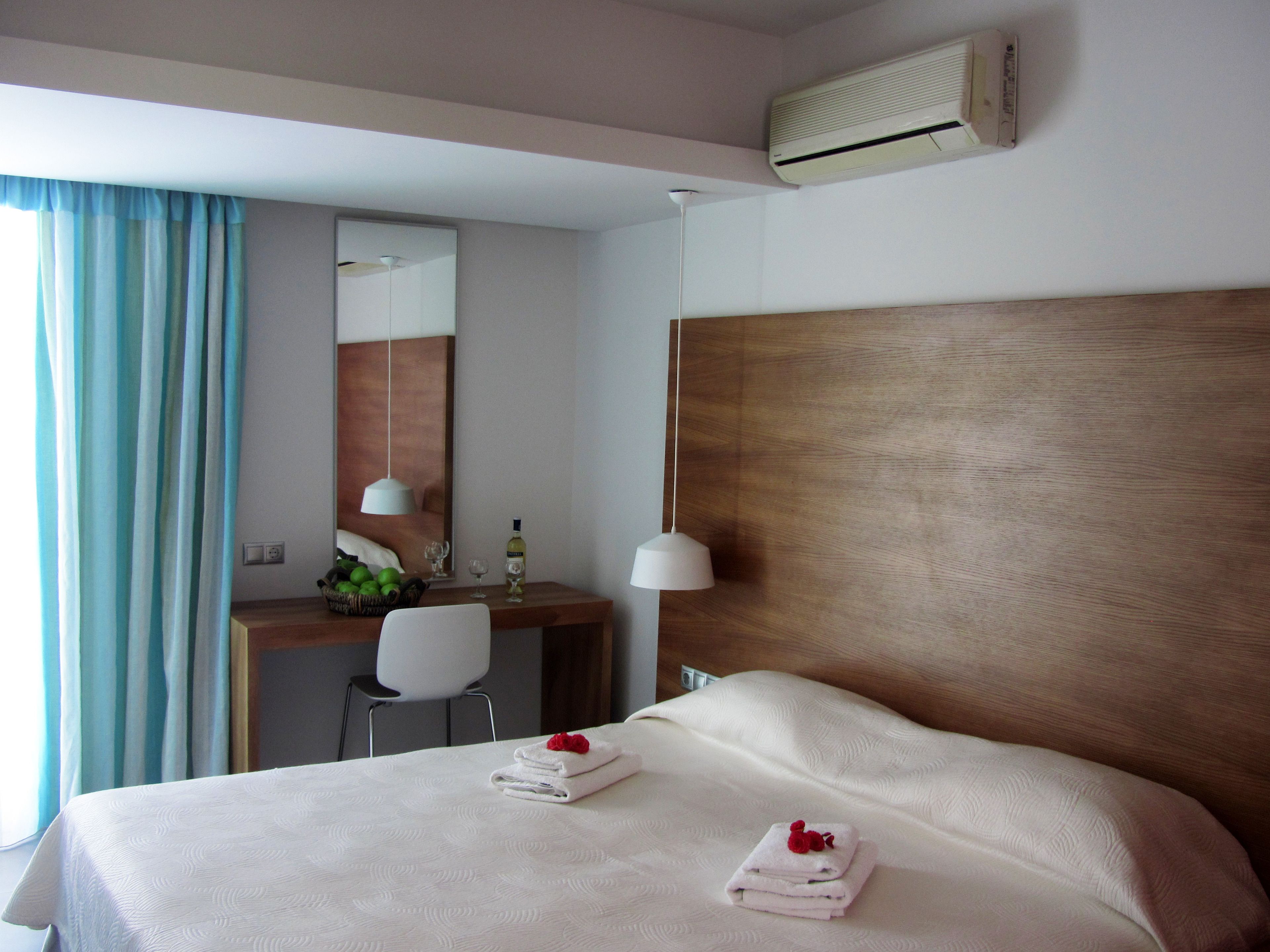 A hotel room with a bed, a desk, a chair, a mirror, and an air conditioning unit.