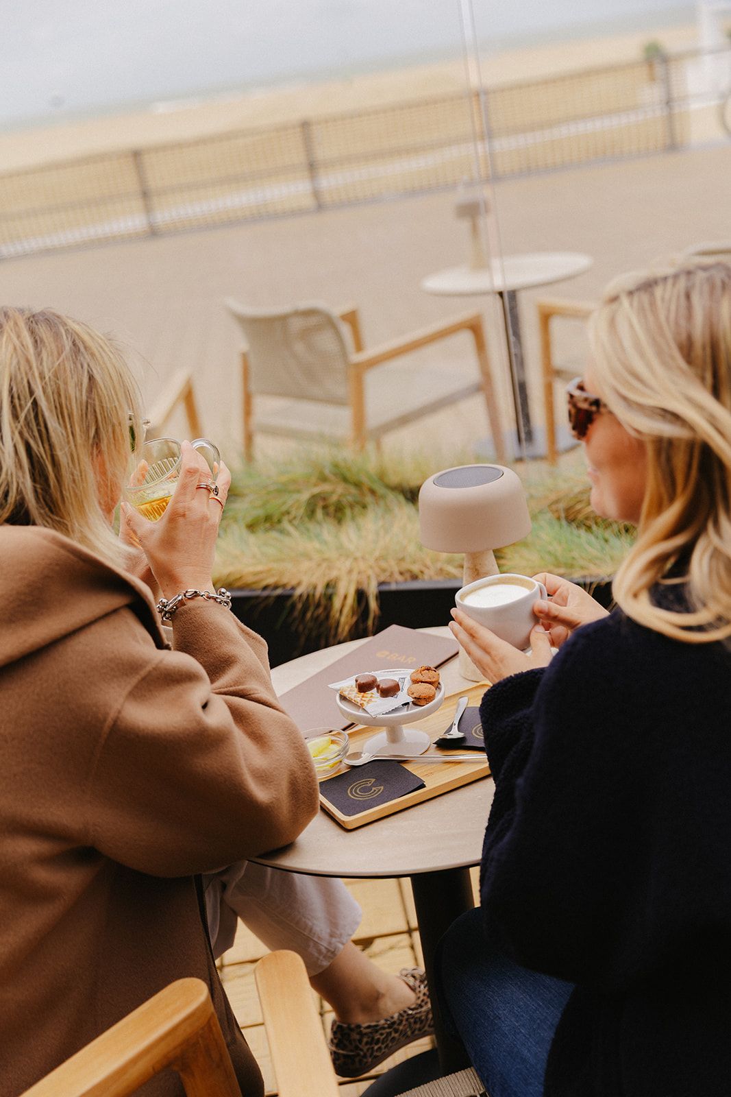 Twee personen zitten aan een buitentafel met een toetjesbord, een kopje en een smartphone.