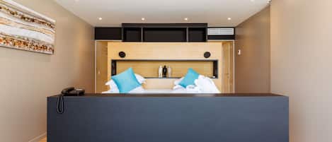 A modern hotel room with a dark reception desk, a wooden headboard, and a large abstract painting on the wall.
