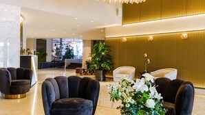 Un hall d’entrée moderne avec un grand lustre, des fauteuils à revêtement sombre, une table basse en verre et un comptoir d’accueil en arrière-plan.