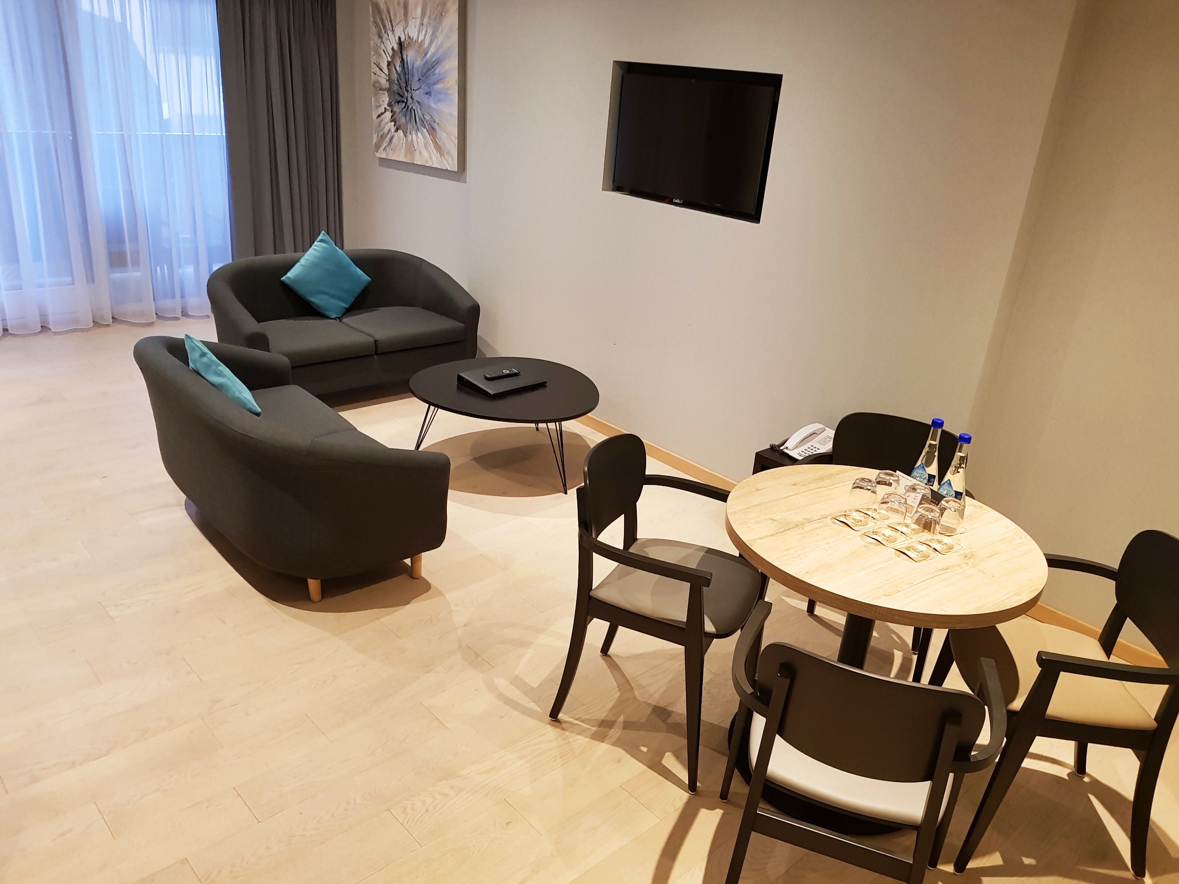 Una sala de estar moderna con un sofá, dos sillones, una mesa de centro y una mesa de comedor con sillas. Hay un televisor de pantalla plana montado en la pared y una ventana con cortinas translúcidas.