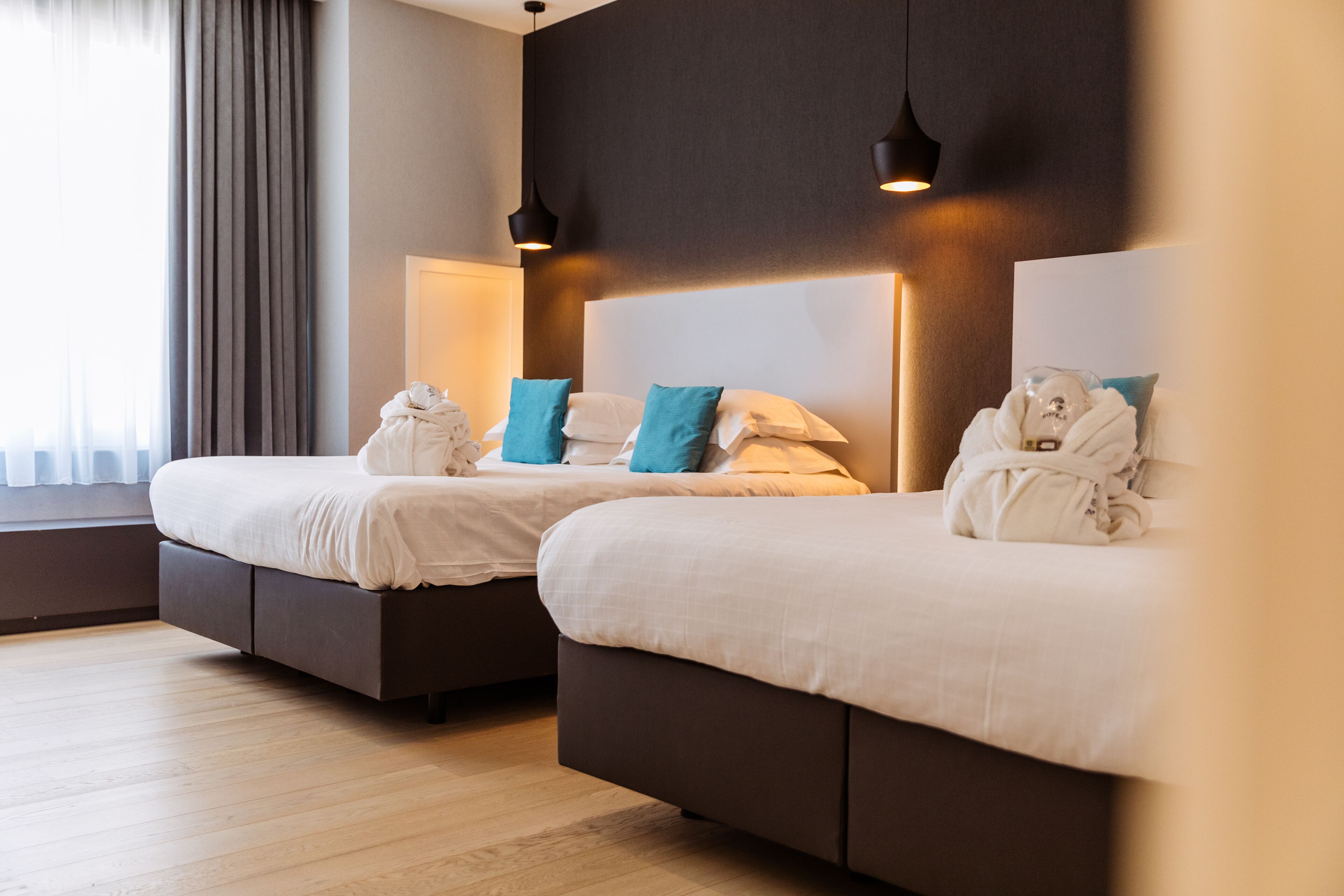 Two beds with white linens and teal pillows, a dark headboard, and a window with curtains.