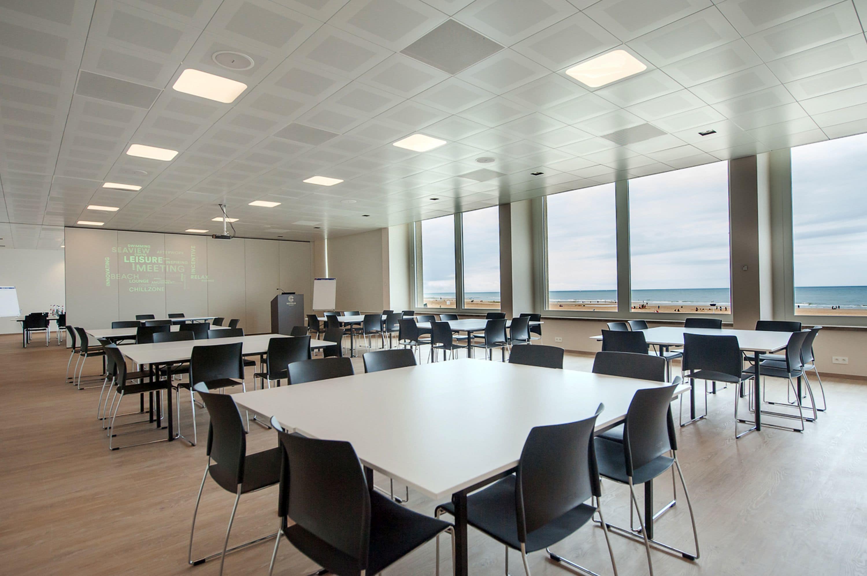 Een vergaderruimte met een grote tafel en stoelen, een projectorscherm en uitzicht op het strand door grote ramen.
