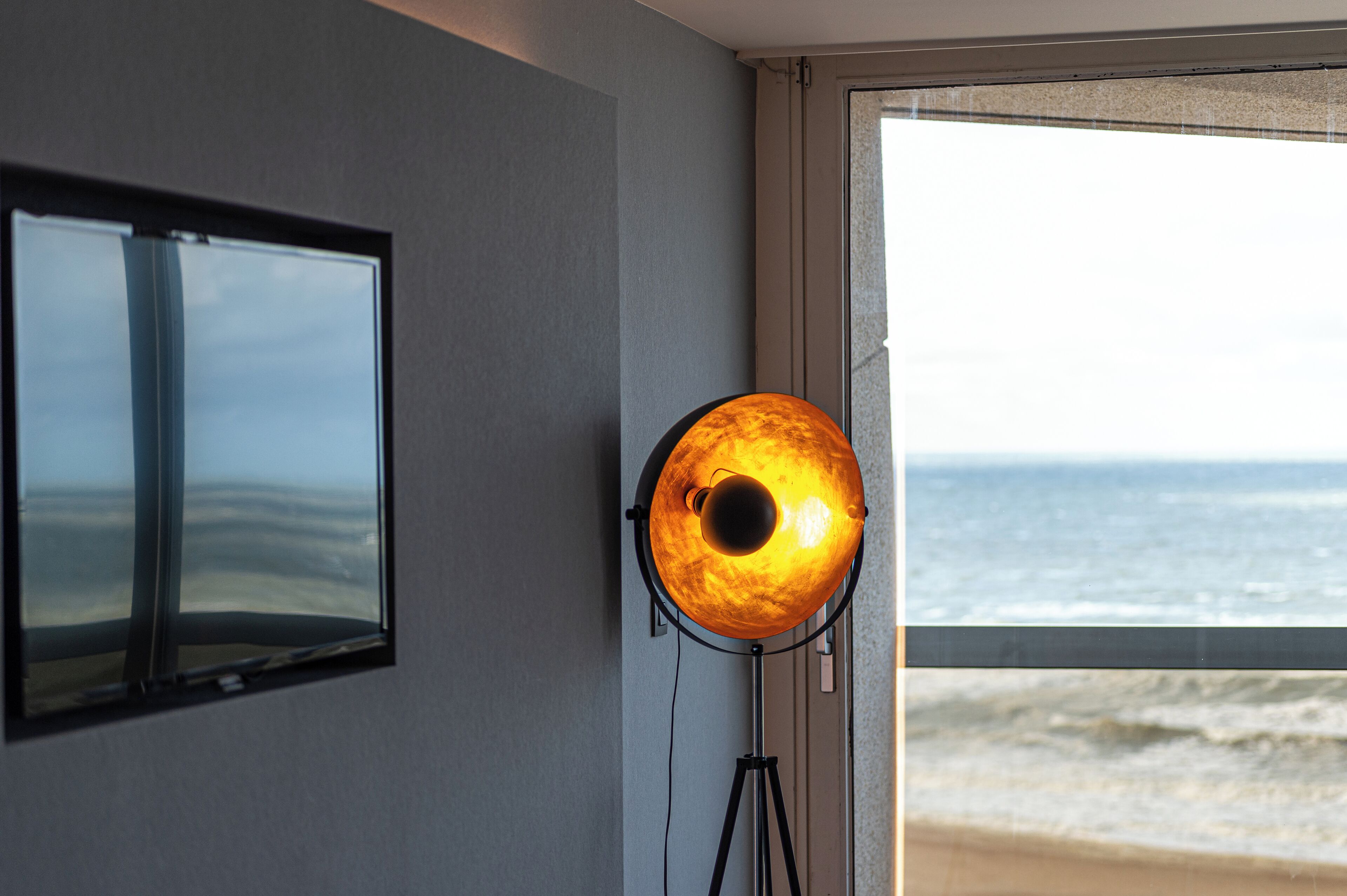 A room with a large window overlooking the beach, a wall-mounted TV, and a floor lamp.