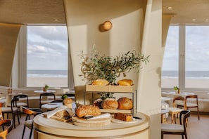 A breakfast buffet with various breads and pastries, a plant centerpiece, and a view of the beach through large windows.