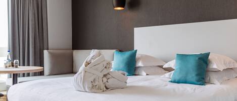 A neatly made bed with white linens and a teal pillow, a round wooden side table with a bottle and glasses, and a dark wall with a hanging light fixture.