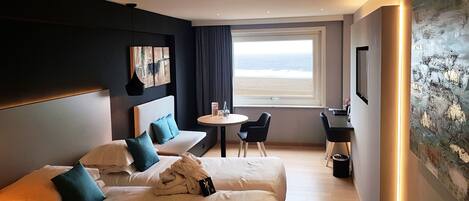 Habitación de hotel con dos camas, un escritorio, una silla, una mesita, un cuadro y un ventanal con vistas a la playa.