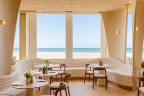 Un restaurante frente al mar con mesas redondas y asientos acolchados, amplias ventanas con vistas al océano.