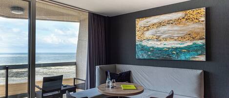 A modern hotel room with a large window overlooking the beach, a round table with chairs, a sofa, and a painting on the wall.