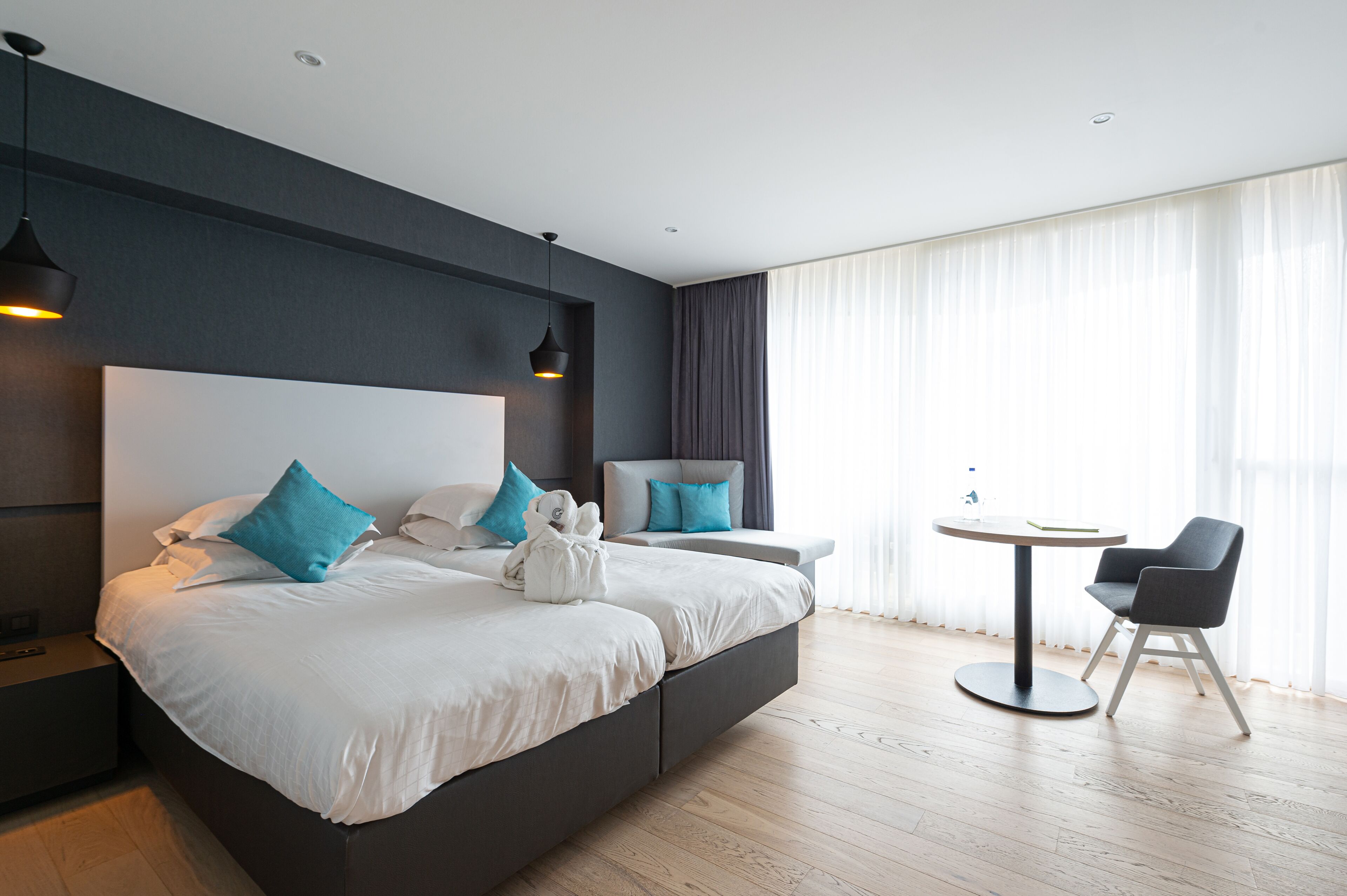 Een moderne hotelkamer met een groot bed, twee nachtkastjes, een ronde tafel met een stoel, en een groot raam met dunne gordijnen.