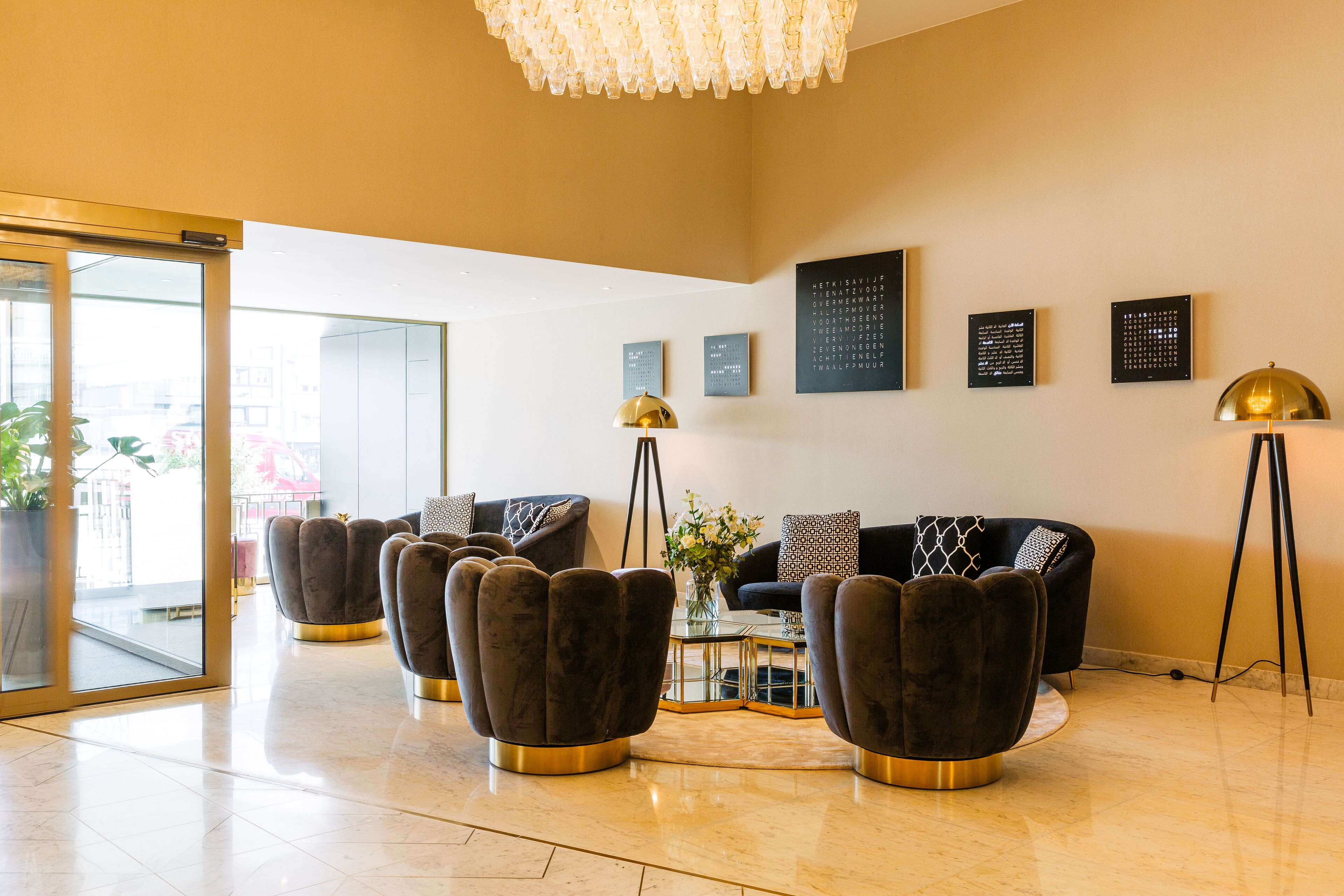 A modern lobby with a seating area featuring dark, tufted chairs with gold bases, a glass coffee table, and a chandelier.