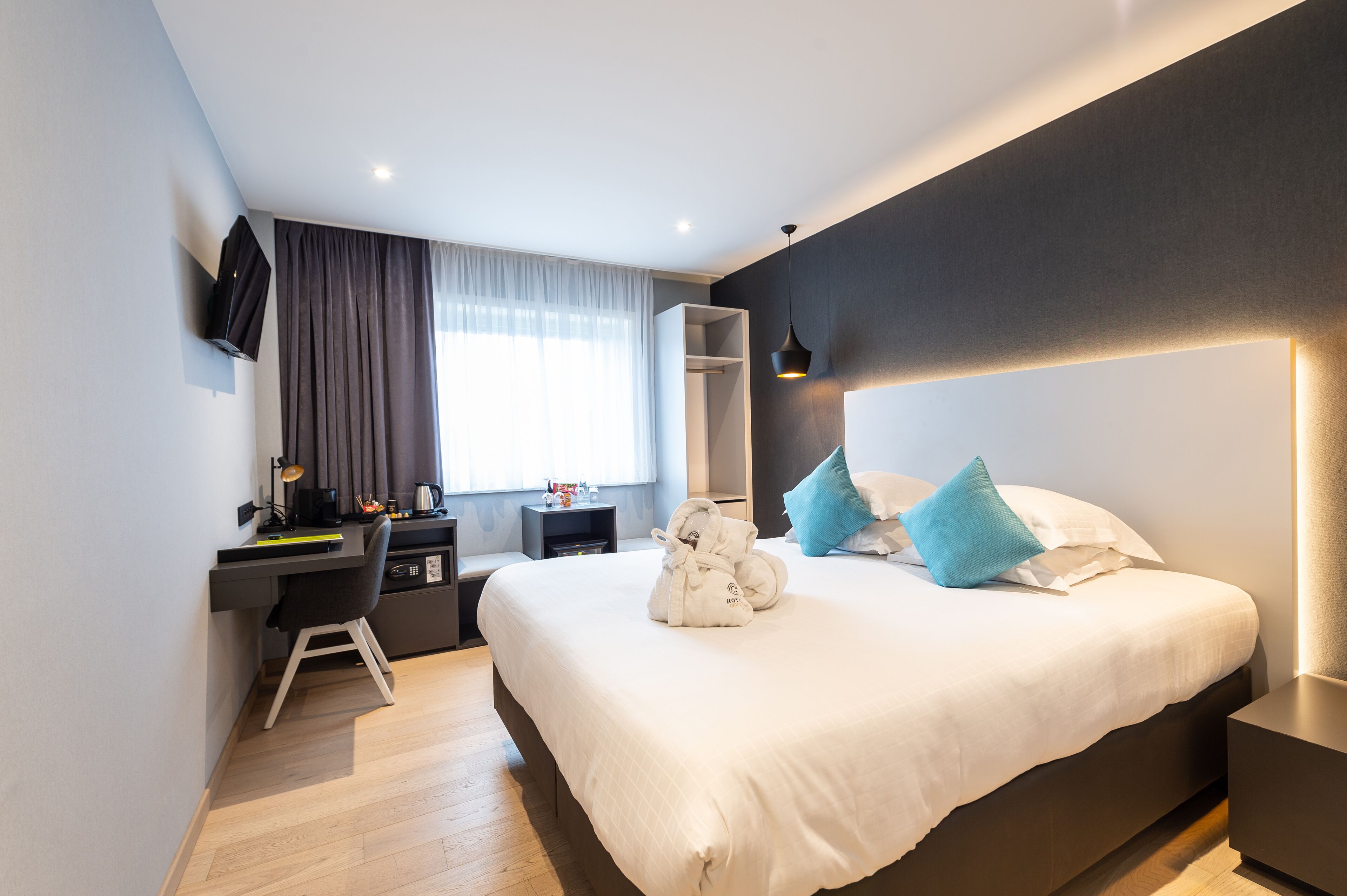 A modern hotel room with a large bed, a desk with a chair, a wardrobe, and a TV mounted on the wall.