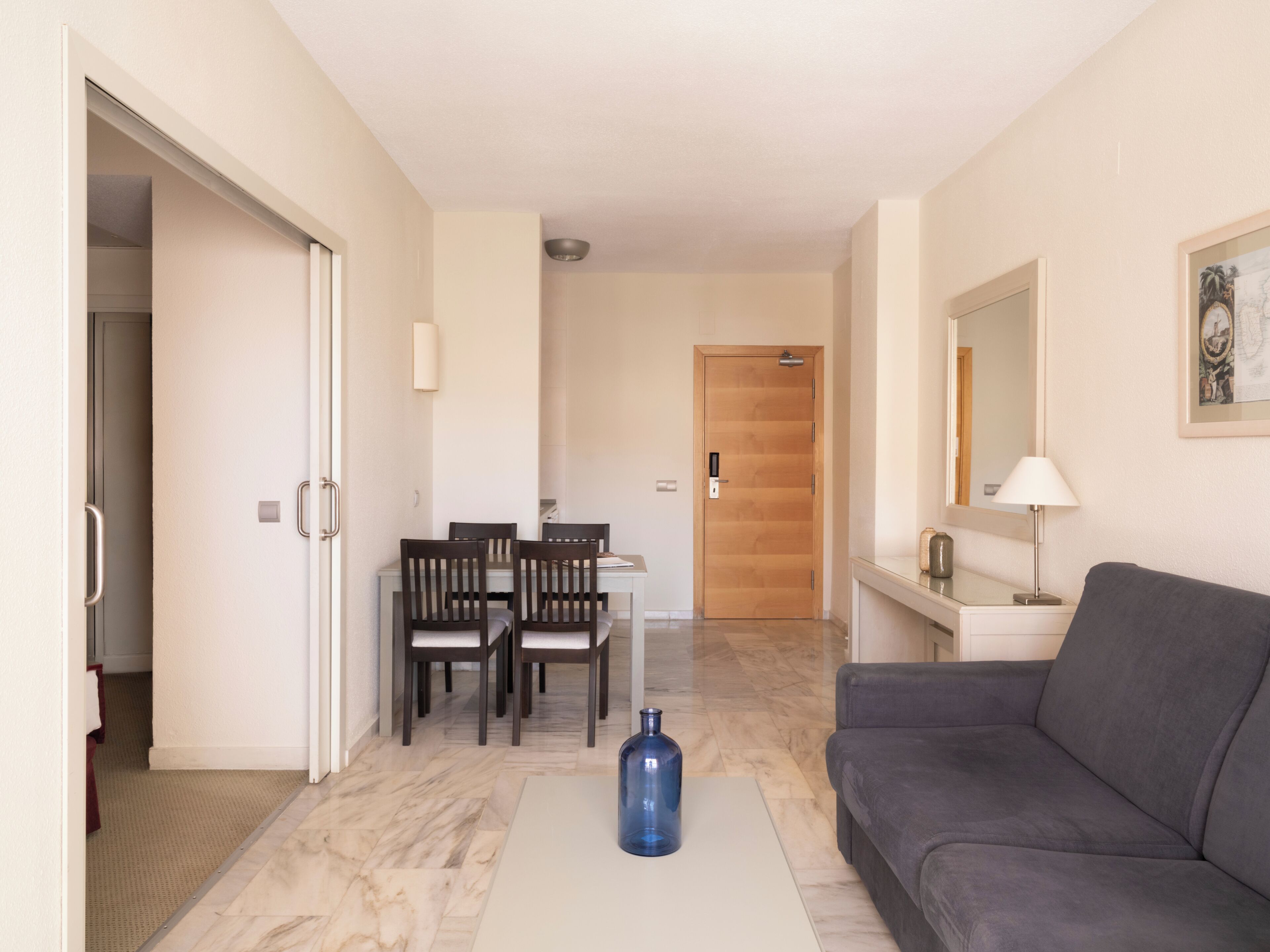 Una sala de estar moderna con un sofá gris, una mesa de comedor con sillas y un jarrón azul sobre una mesa de centro blanca.
