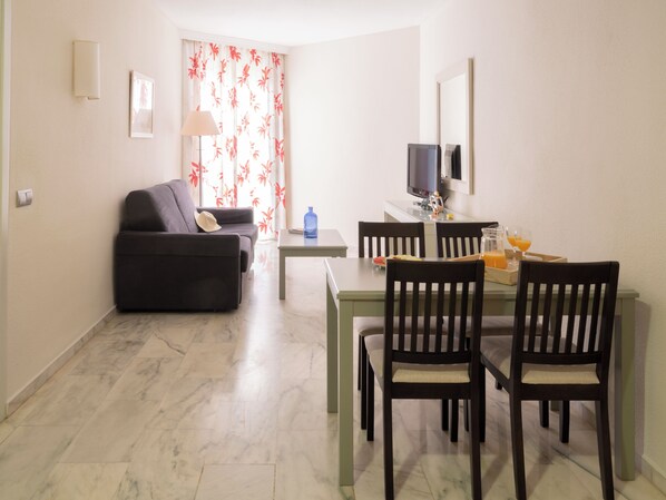 A living room with a sofa, a dining table with chairs, a TV, and a window with floral curtains. - Ona Marinas de Nerja Spa Resort (Nerja)