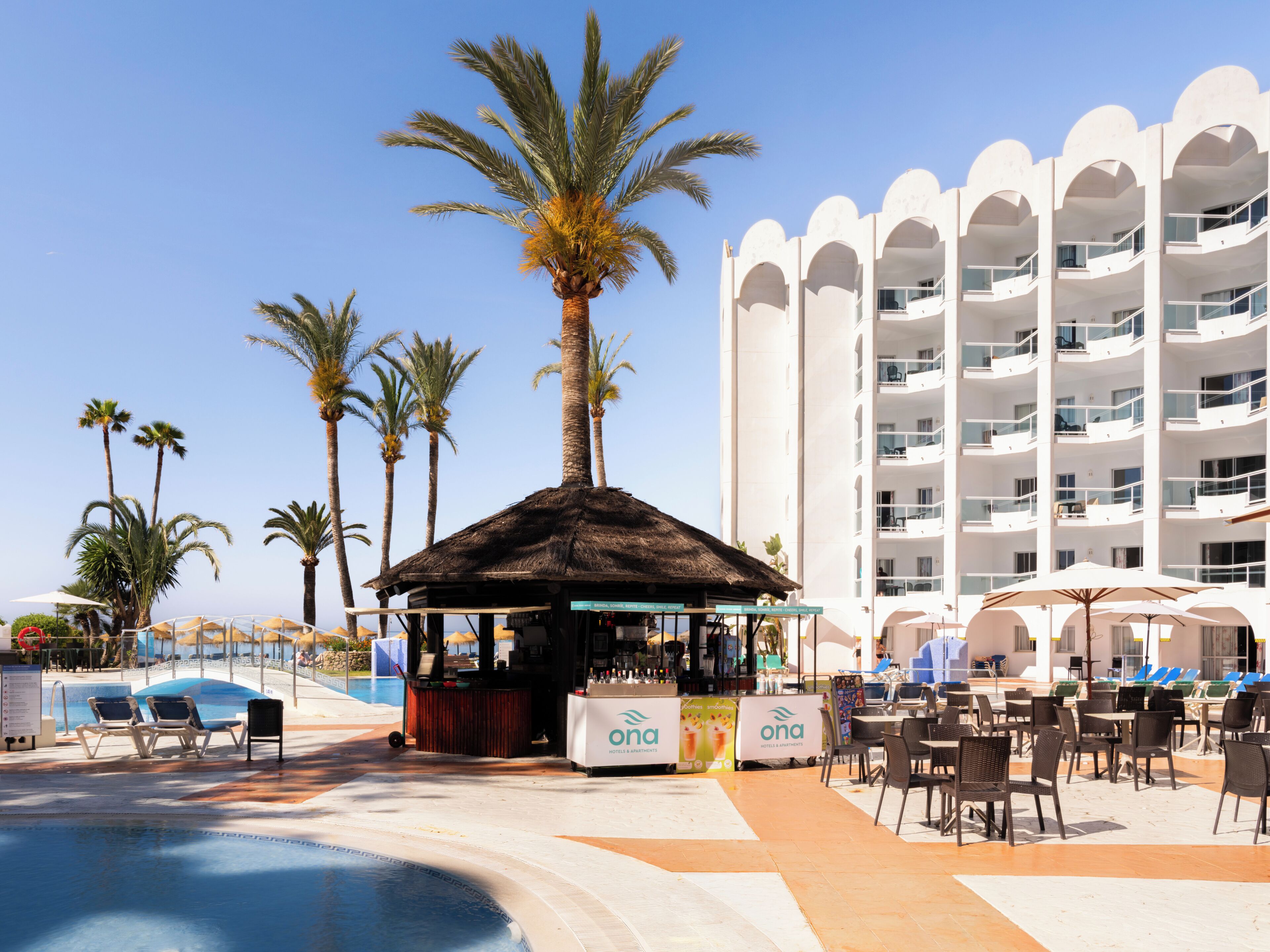 Un área de piscina en un resort, con sillones de descanso, un bar y un edificio con balcones.
