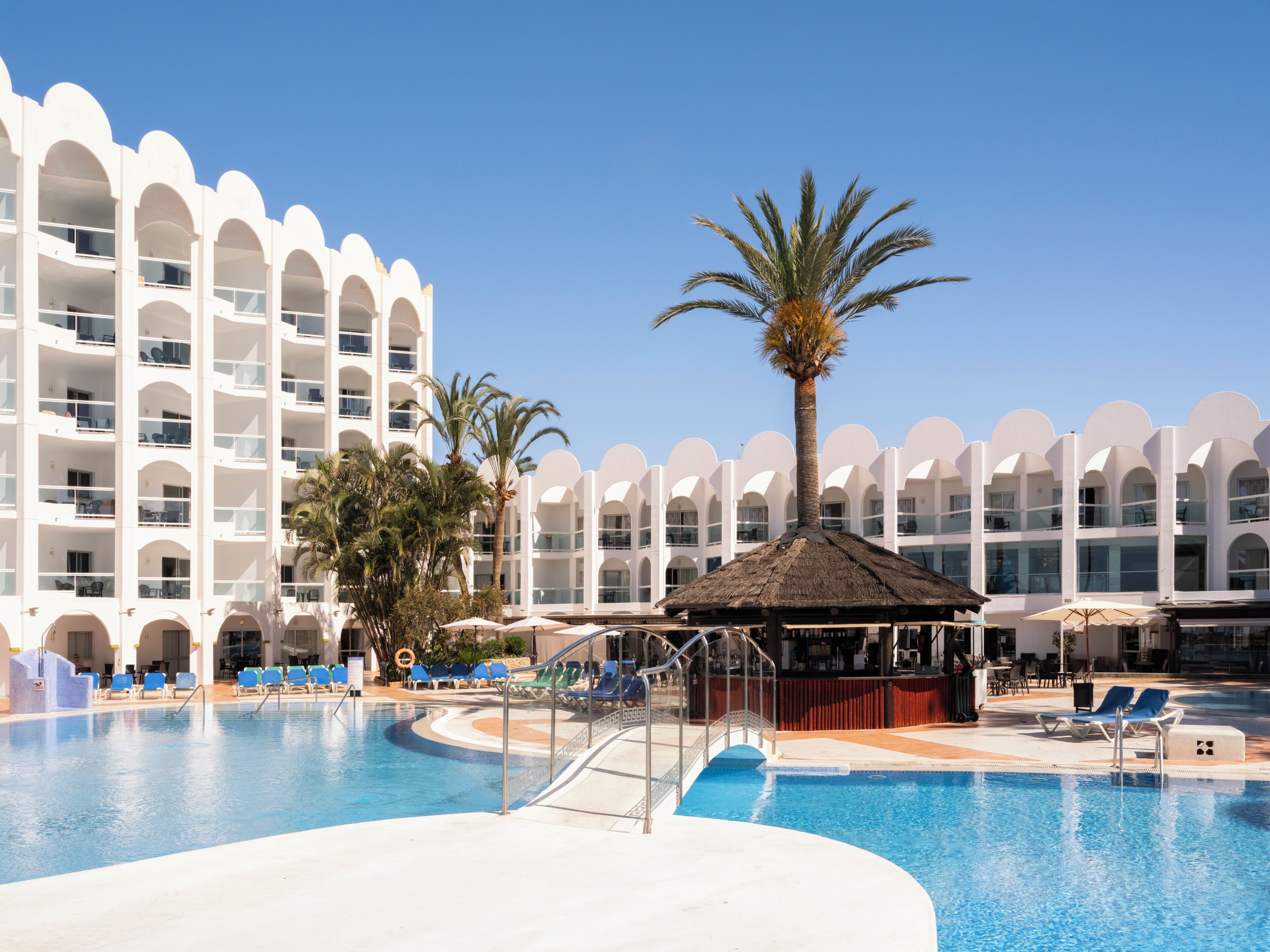 Et hotel med swimmingpool, palmer og en struktur med stråtæthed.