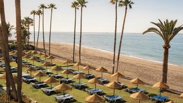 A beachside resort with rows of sun loungers and thatched umbrellas, palm trees, and a view of the ocean.