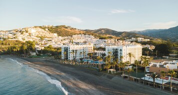A coastal hotel with multiple white buildings, a beachfront, and palm trees.