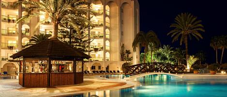 Zona de piscina nocturna en un resort, con bar, sillones de descanso y palmeras.