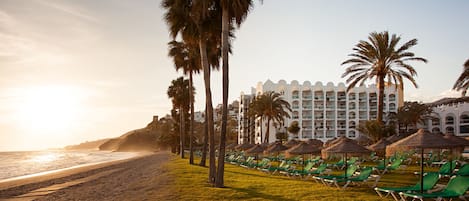 A beachfront hotel with rows of sun loungers, palm trees, and a large building with multiple balconies.