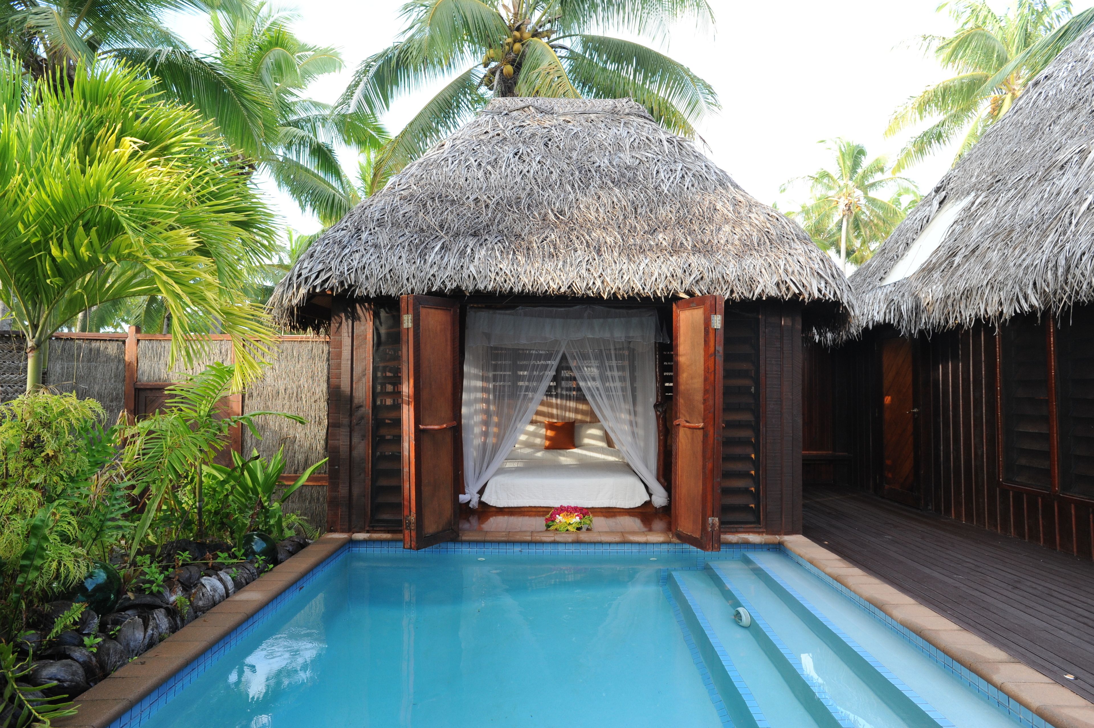 A thatched-roof hut with a bed, a pool, and lush tropical vegetation.