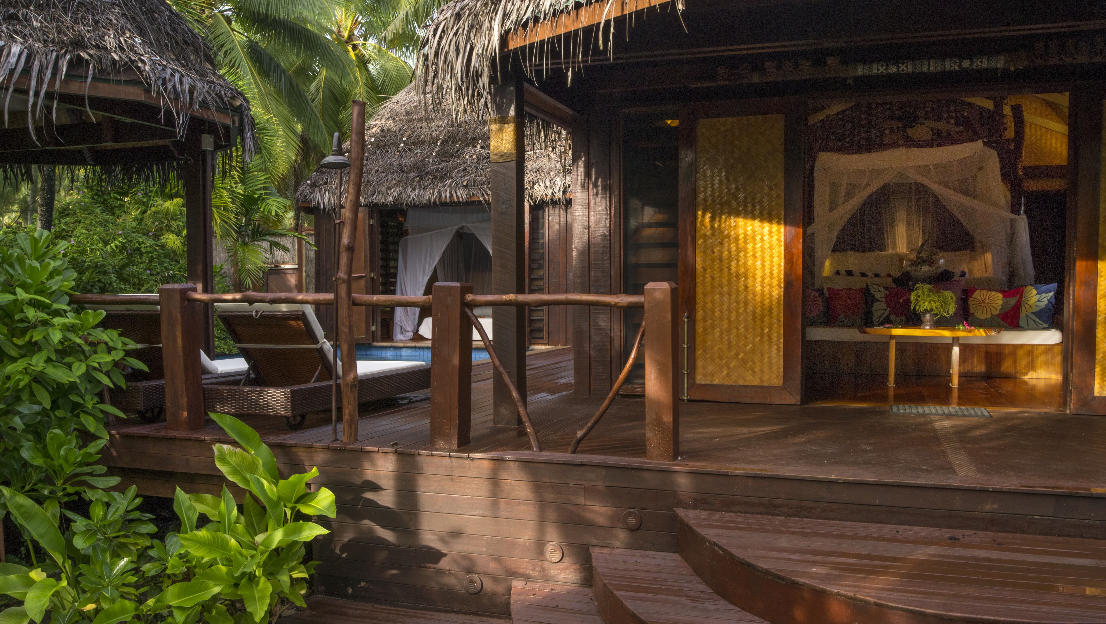 A wooden deck with a thatched-roof structure, a seating area with cushions, and a small table with a plant.