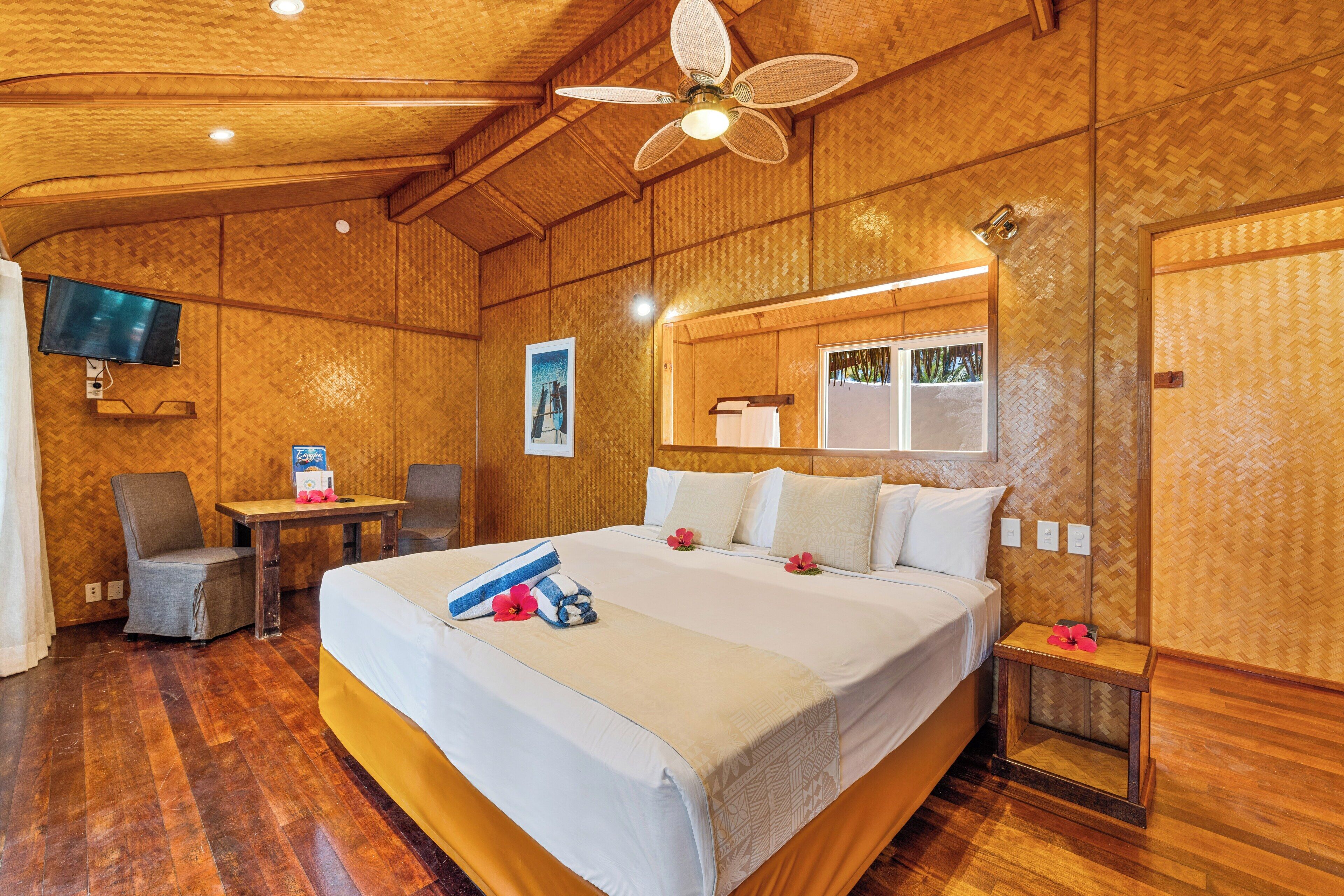A hotel room with a wooden interior, a bed with white bedding, a desk with a chair, a television on the wall, and a ceiling fan.