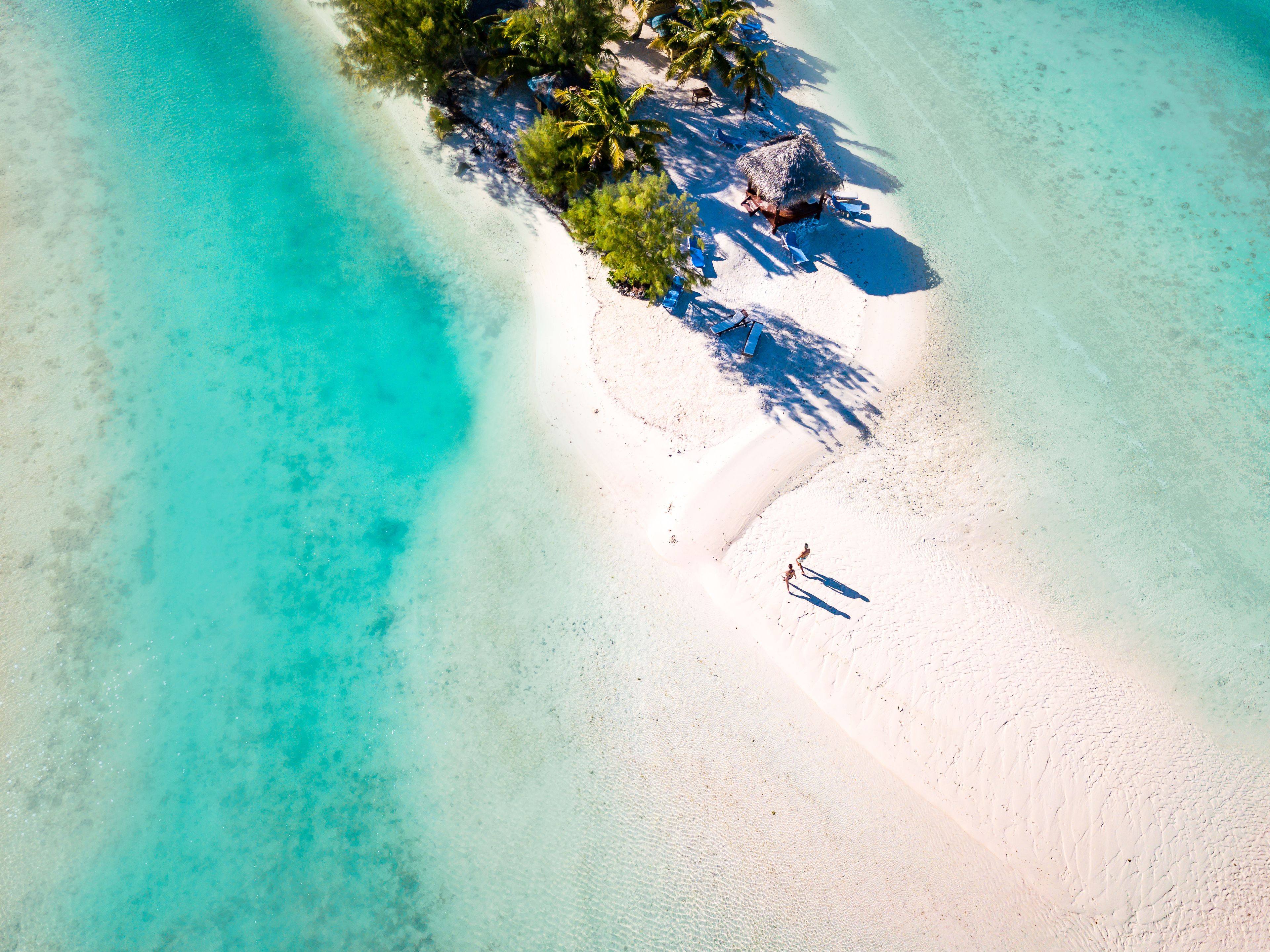 Een luchtbeeld van een strand met helder turquoise water, wit zand en enkele palmbomen.