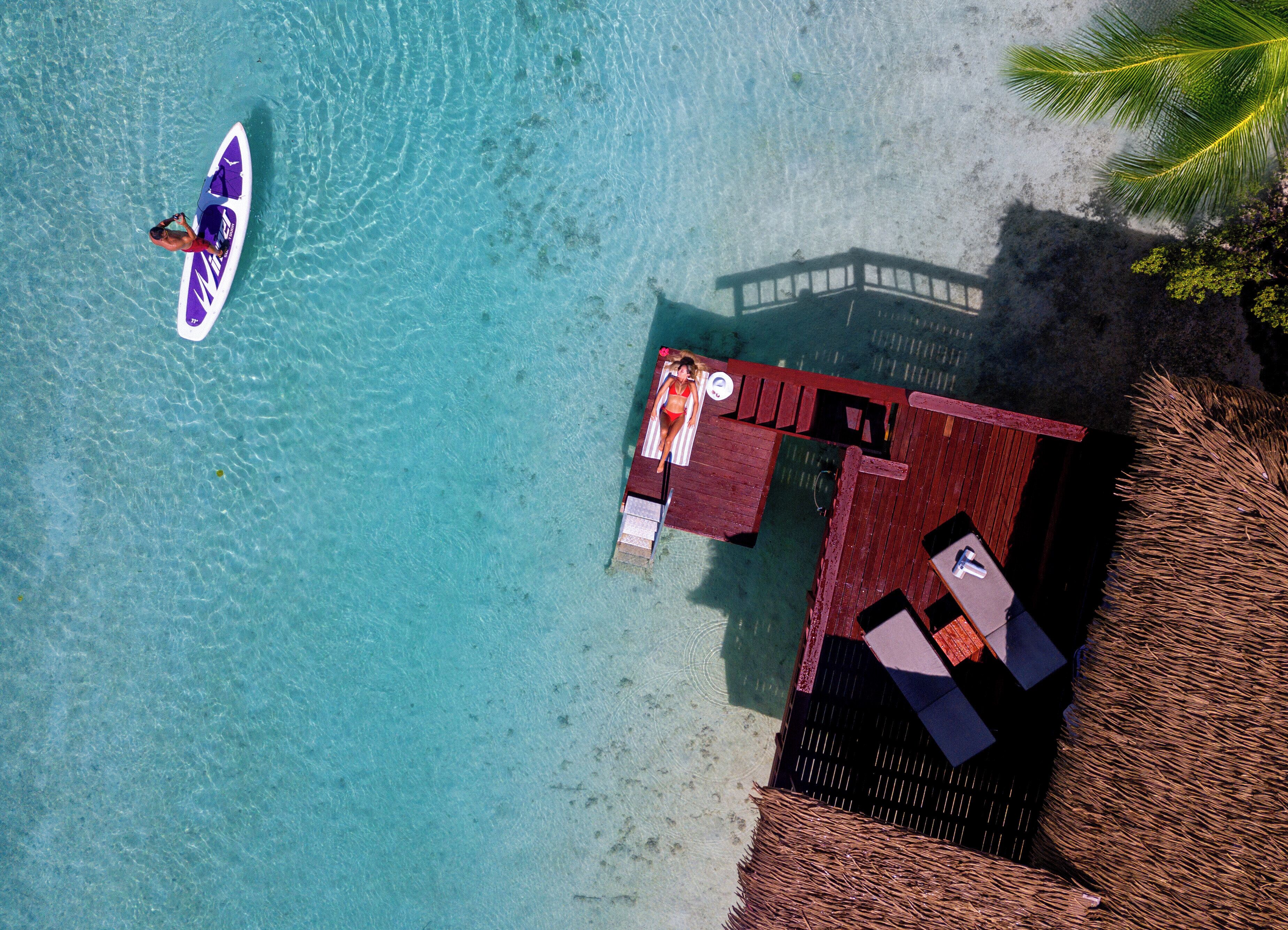 An aerial view of a beachside resort with a clear turquoise sea, a wooden deck with lounge chairs, and a thatched-roof structure.