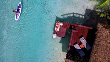 An aerial view of a beachside resort with a clear turquoise sea, a wooden deck with lounge chairs, and a thatched-roof structure.
