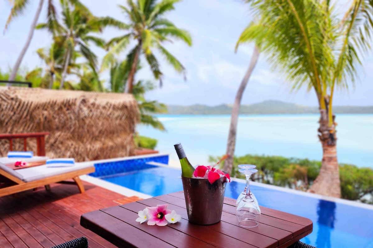 A poolside area with lounge chairs, a table with a bottle of wine and glasses, and a view of the ocean.