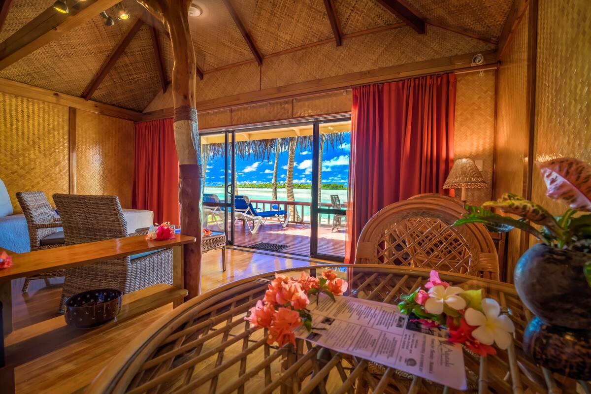 A room with a wicker sofa, a dining table with a menu, and a view of the beach through large windows.