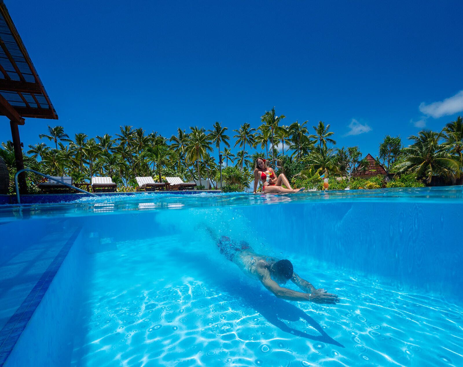 A swimming pool with clear blue water, surrounded by palm trees and lounge chairs.