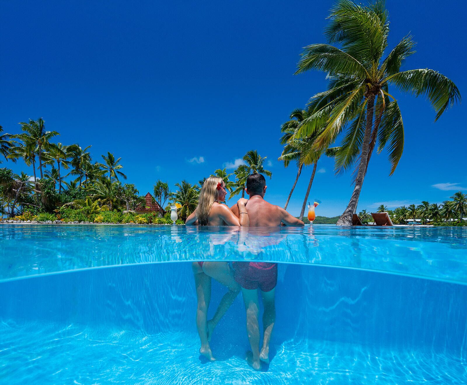 Una parella en una piscina cristal·lina amb un paisatge tropical de palmeres i cel serè.